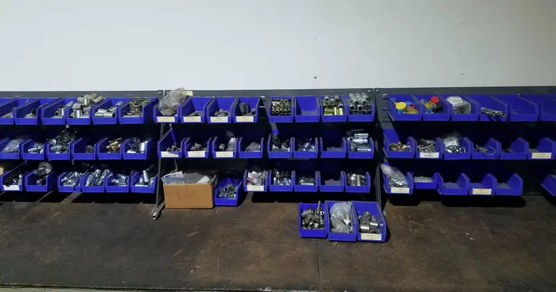 Three rows of blue plastic bins on a wall rail, each holding steel and brass A/C hose fittings, ferrules and caps, with printed labels