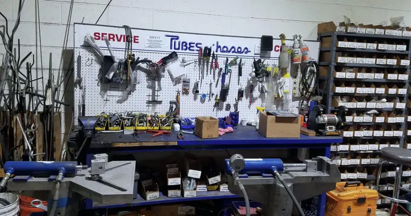 A blue steel workbench under a white pegboard hung with pliers, cutters and crimp dies, a printed Tubes n’ hoses sign above it, two hydraulic crimpers in the foreground and a rack of small parts boxes on the right