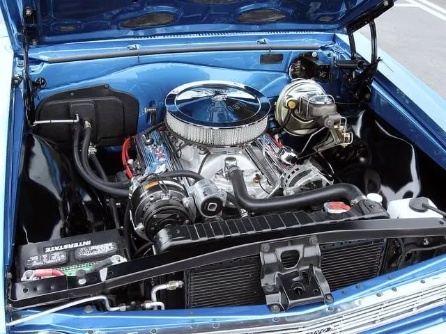 The engine bay of a metallic blue 1960s Chevrolet with the hood up: a chrome air cleaner on a V8, a chrome A/C compressor and brake booster, a battery at the left