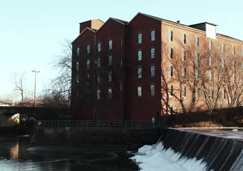 A tall brick mill beside a river dam in winter light, bare trees around it and a concrete bridge behind