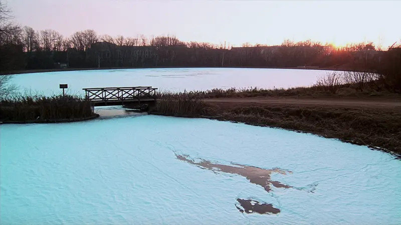 A frozen millpond at sunrise, a small wooden footbridge over the outlet, bare trees on the far shore