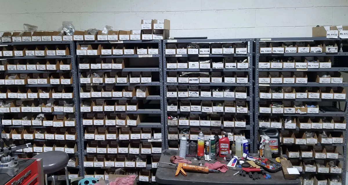Four bays of grey steel shelving packed with small labelled parts boxes, a work cart with spray cans and rags in front, a red tool chest at the left