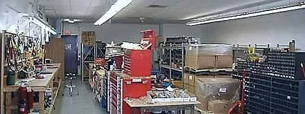 A long shop room under fluorescent tubes: a wooden workbench with a vise on the left, a red tool chest, grey shelving with boxes and crates on the right