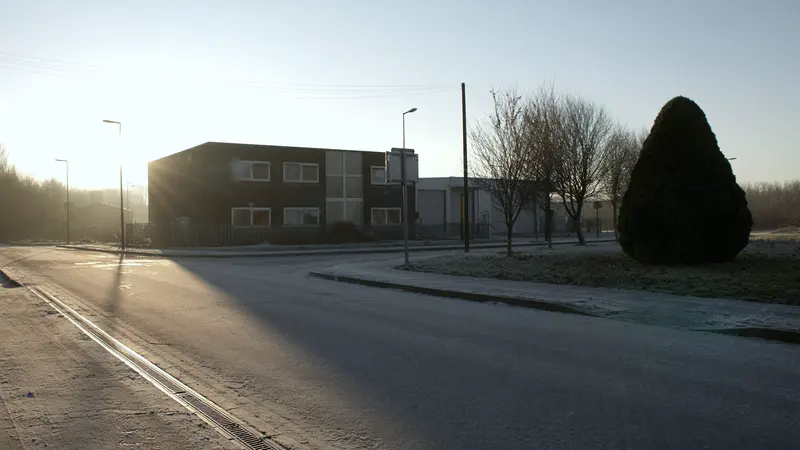 A quiet light industrial street on a frosty morning, low sun, a dark two storey office building with a fenced yard, a taller warehouse behind, bare trees and a conifer