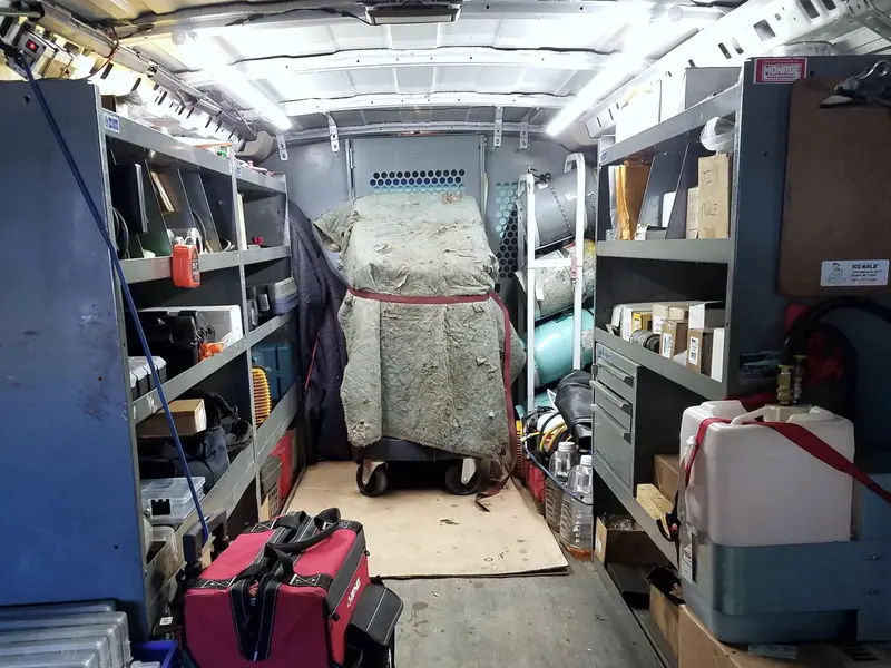 The cargo bay of a service van from the open rear doors: steel shelving with parts bins down both sides, a red tool bag on the floor, a machine under a moving blanket strapped to a cart at the far end